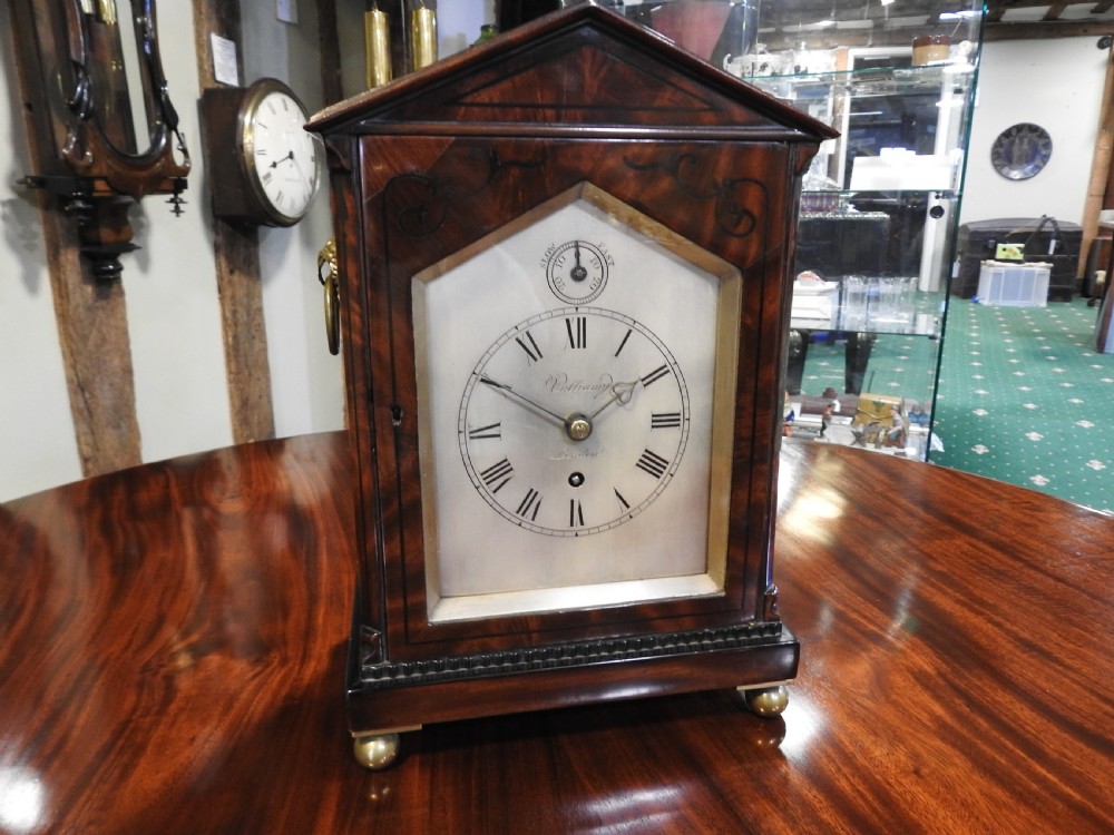 An antique Regency flame mahogany bracket clock featuring a gabled top, a silvered dial signed Vulliamy London, and brass ball feet.