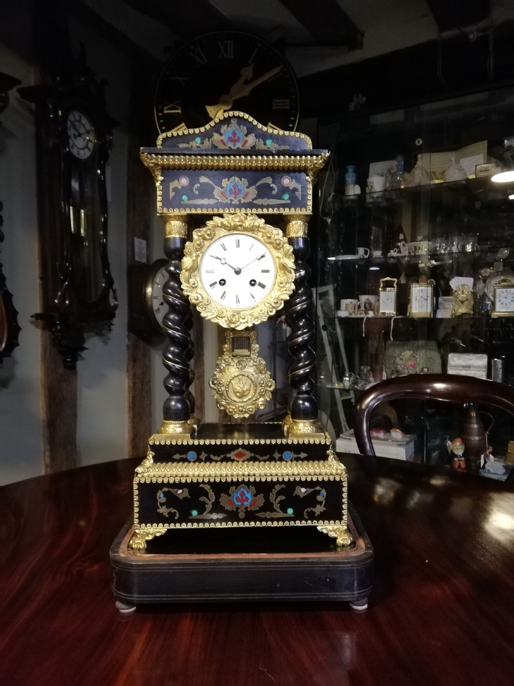 A 19th-century French portico mantel clock featuring ebonised barley twist columns, gilt bronze mounts, stone inlay, and an original glass cloche. View XV