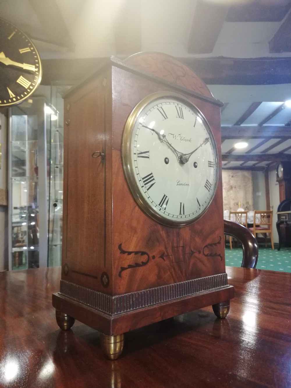An antique mahogany bracket clock featuring an arched top, inlaid woodwork, a white dial with Roman numerals signed W. Clark London, and brass bun feet. View VIII