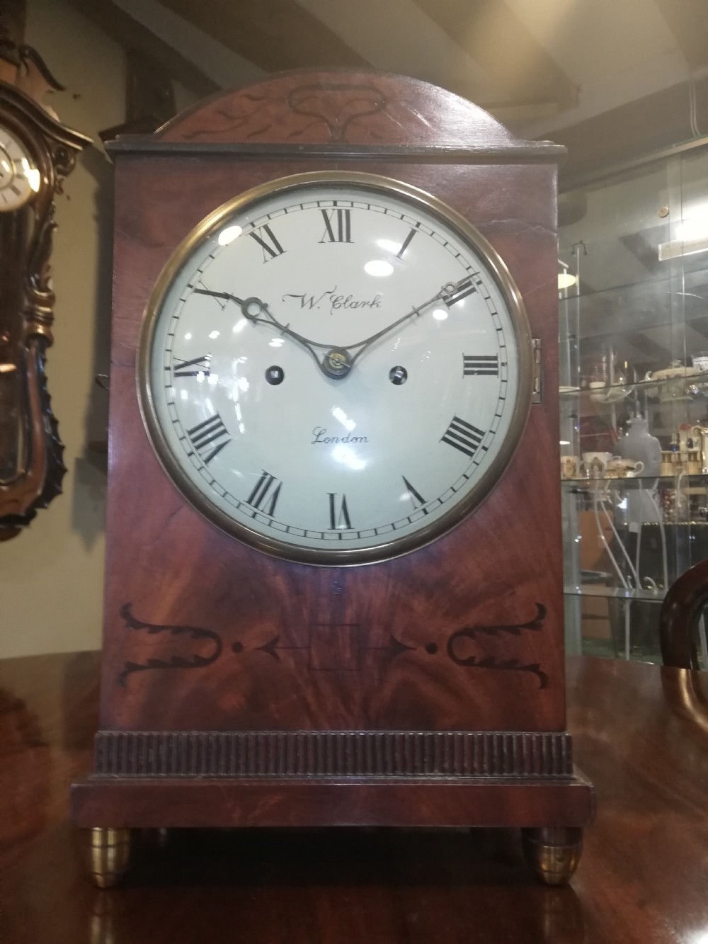 An antique mahogany bracket clock featuring an arched top, inlaid woodwork, a white dial with Roman numerals signed W. Clark London, and brass bun feet.