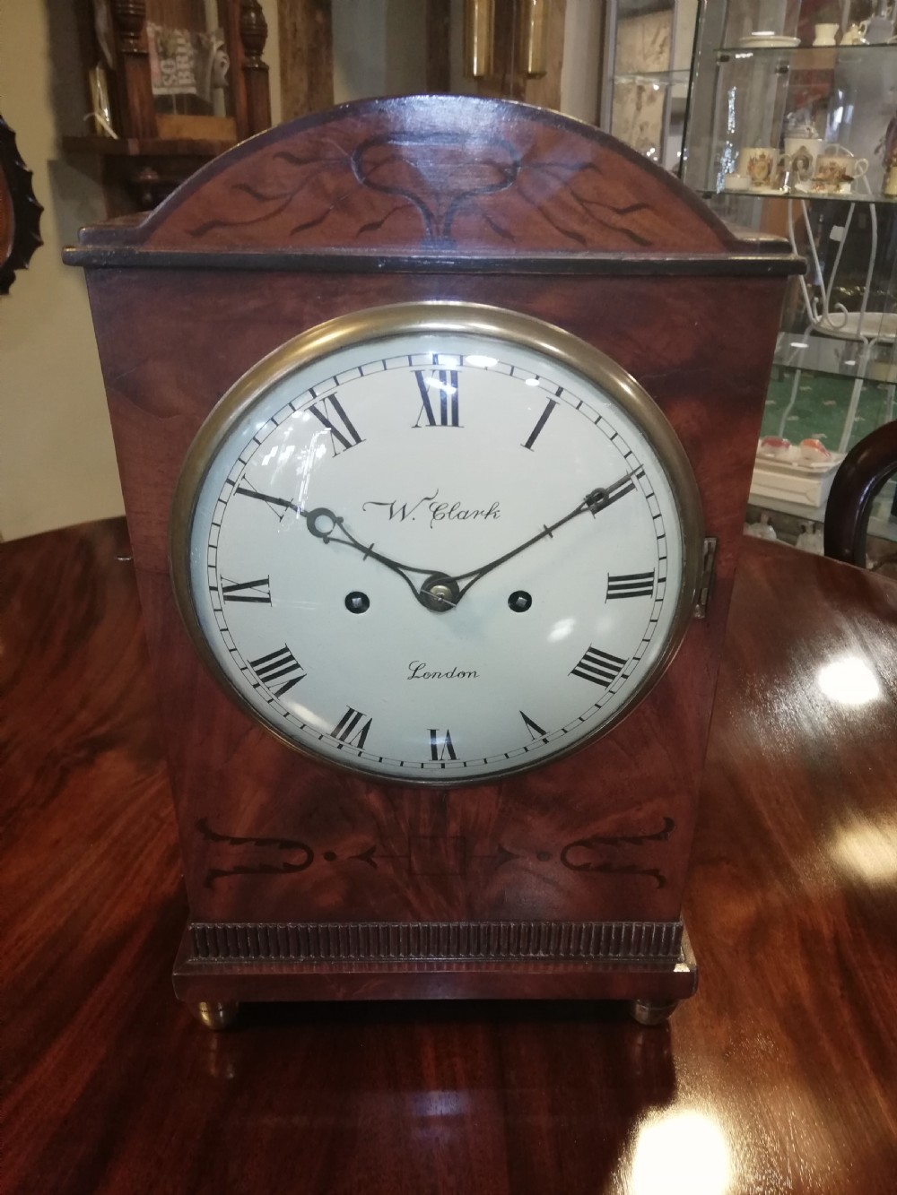 An antique mahogany bracket clock featuring an arched top, inlaid woodwork, a white dial with Roman numerals signed W. Clark London, and brass bun feet. View XI