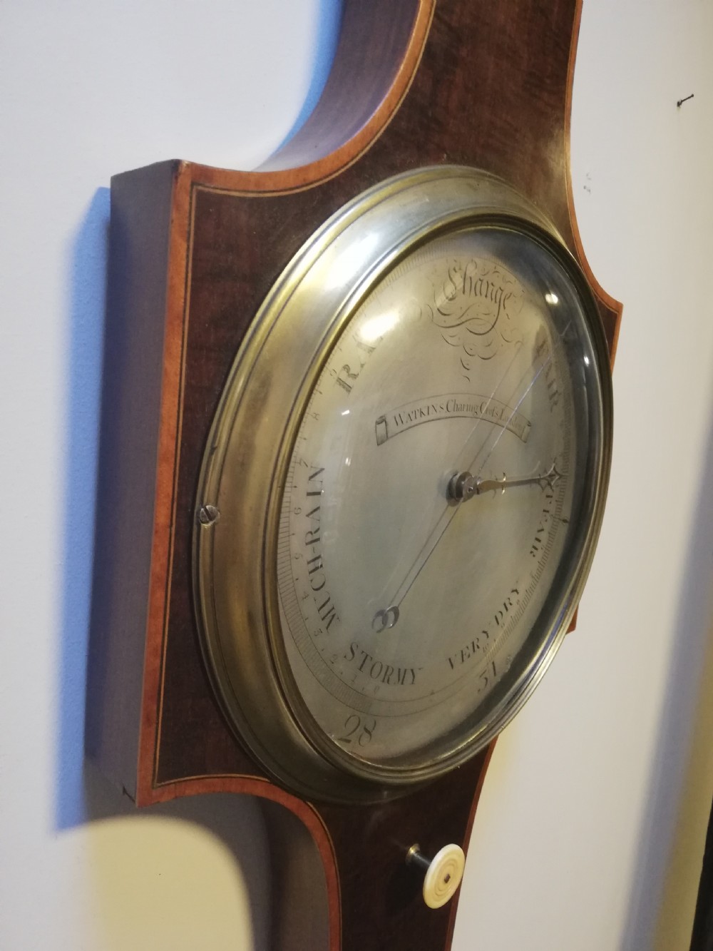An antique Georgian mahogany wheel barometer featuring a silvered dial with brass bezel, dark wooden casing with inlay, and a pediment top. View XVIII