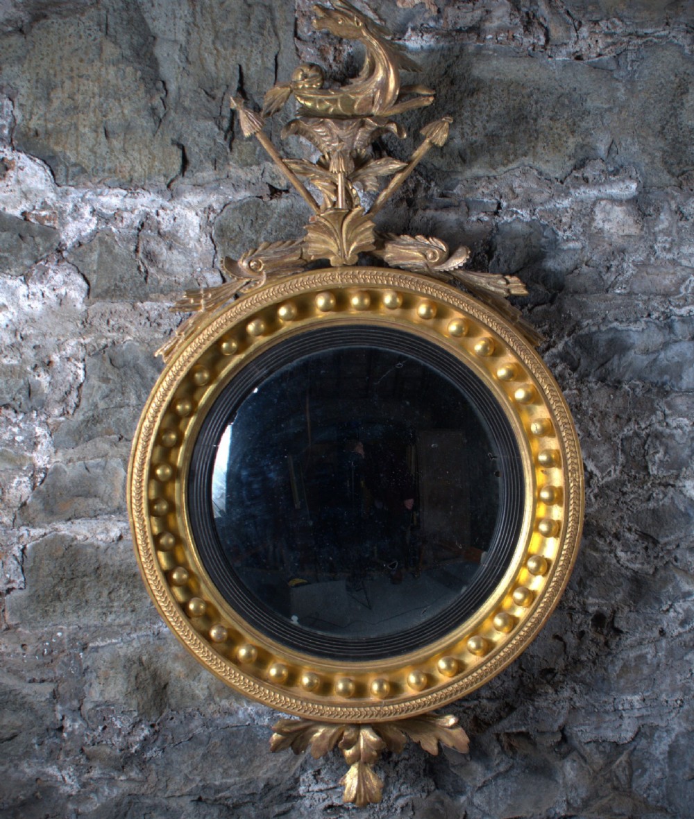 A circular Regency giltwood convex mirror featuring a carved dragon crest atop a moulded frame adorned with spherules and an acanthus leaf base pendant. View VI