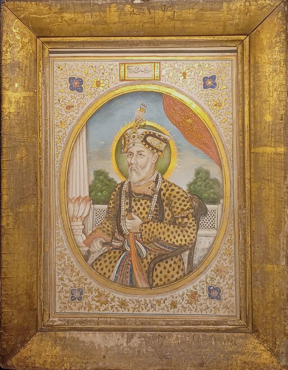 An oval portrait miniature painting of Mughal Emperor Akbar II wearing a jewelled turban, set within an illuminated floral border and a gilded wooden frame.