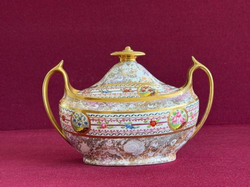fig 1. An oval porcelain sucrier with a lid, twin handles, gilded accents, and hand-painted floral roundels on a detailed ground.