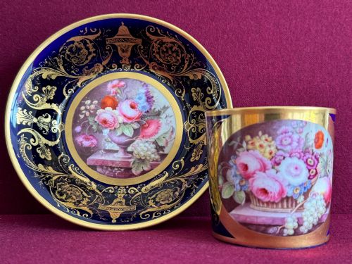 fig 1. A cylindrical porcelain coffee can and saucer featuring deep cobalt blue grounds, elaborate gilt scrollwork, and central hand-painted floral still-life panels.