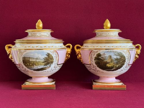 fig 1. A pair of antique Regency porcelain ice pails featuring pink and white grounds, hand-painted landscape scenes, gilded lion mask handles, and pinecone finials.
