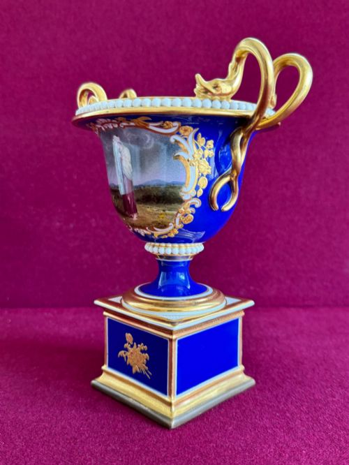 fig 1. A Regency cobalt blue and gilded porcelain urn featuring a central hand-painted panel of a standing figure in a landscape, twin scroll handles, beaded borders, and a square pedestal base.