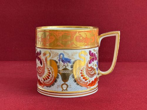 fig 1. A cylindrical Regency period Derby porcelain porter mug decorated with hand-painted bird motifs, gold leaf foliage, an orange top border, and an angular gilded handle.