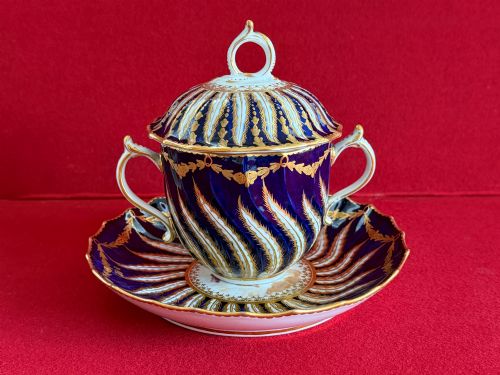 fig 1. An 18th-century cobalt blue and gilded soft-paste porcelain chocolate cup with lid alongside a matching saucer decorated with a painted central cherub motif.