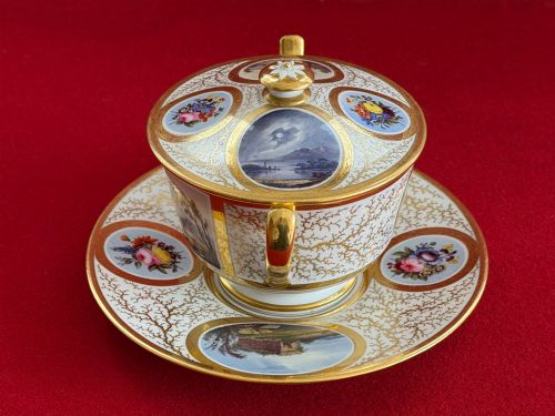 fig 1. An antique Flight, Barr and Barr Worcester porcelain covered chocolate cup with two handles and matching saucer, decorated with hand-painted landscape and floral panels amid gold vermicelli detailing.