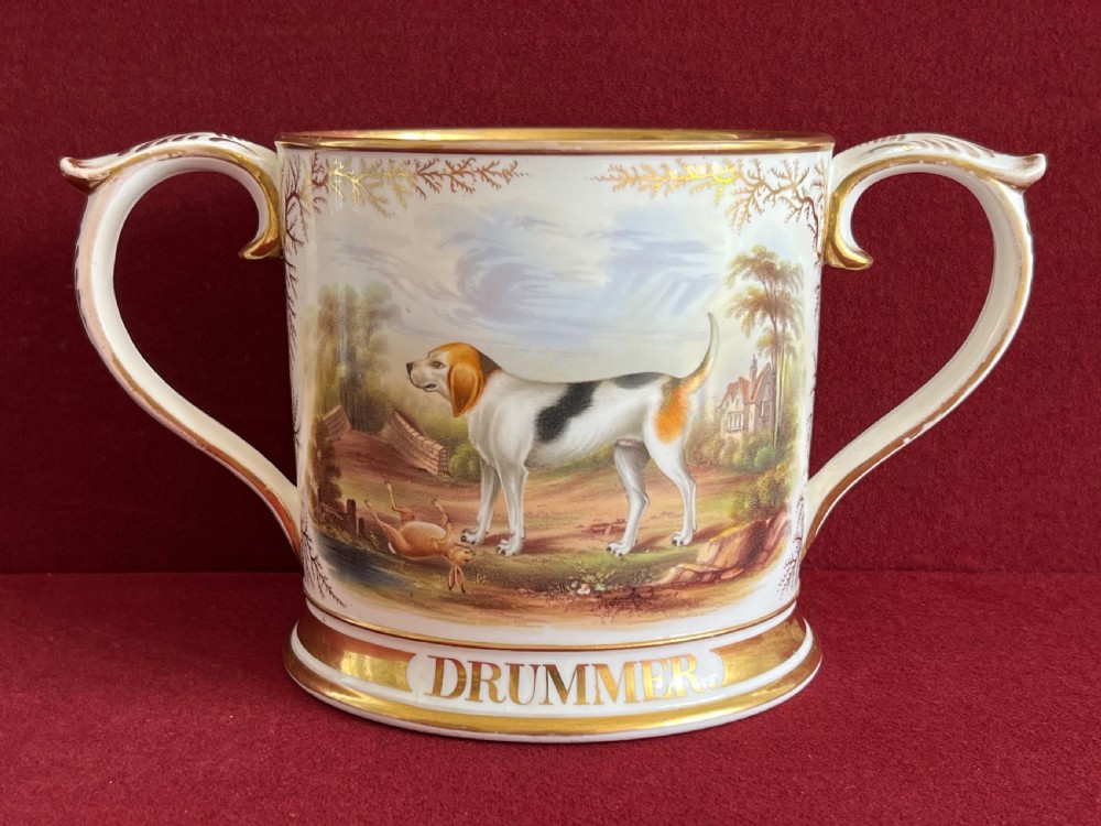 A Victorian English porcelain loving cup featuring two gilded scroll handles and a hand-painted hound named Drummer in a rural setting.
