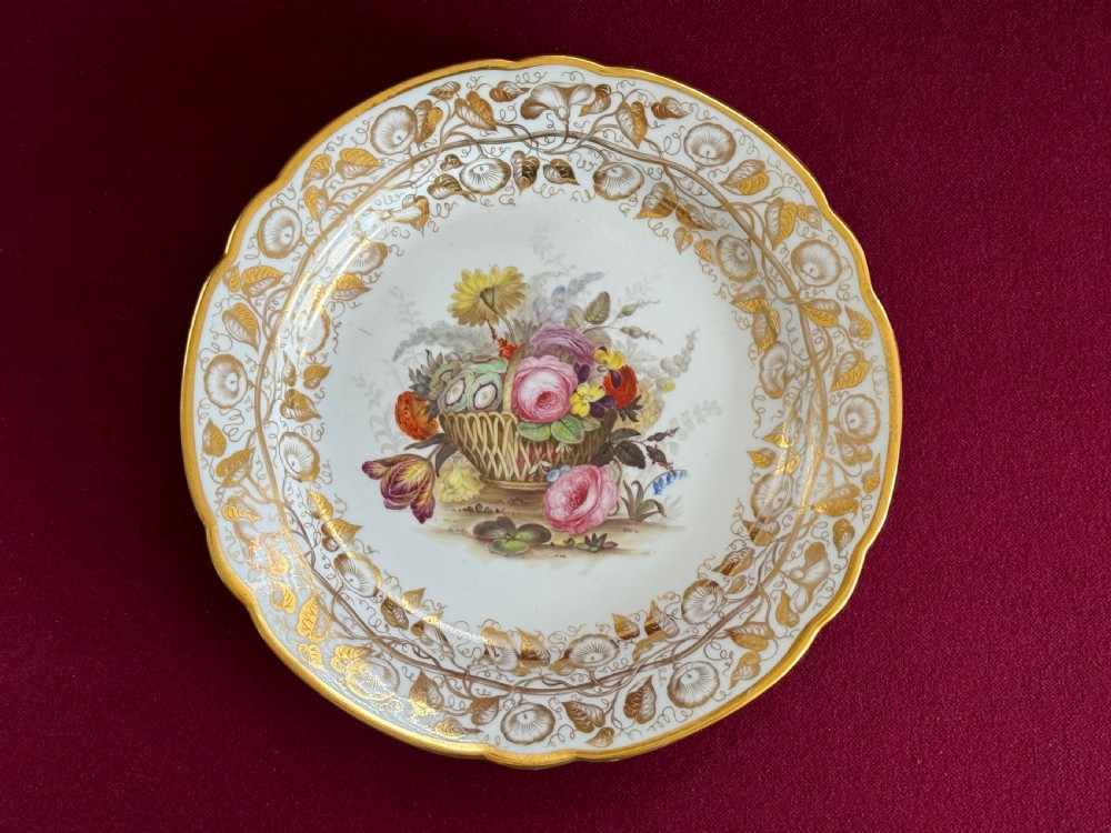 A Regency era circular porcelain plate featuring a scalloped rim, a gold leaf foliate border, and a hand-painted central basket of colourful flowers.