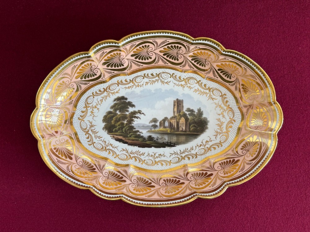 An oval Regency Worcester porcelain dessert dish featuring a hand-painted central river landscape with ruins, bordered by elaborate gold anthemion patterns over a pink background and a scalloped rim.