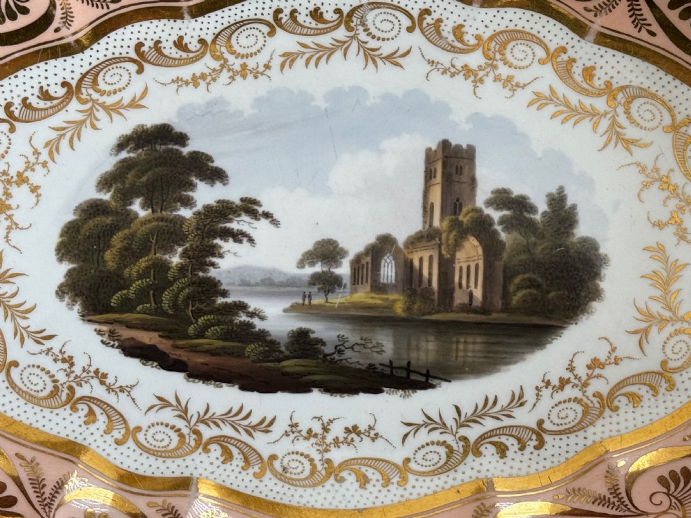 An oval Regency Worcester porcelain dessert dish featuring a hand-painted central river landscape with ruins, bordered by elaborate gold anthemion patterns over a pink background and a scalloped rim. View III