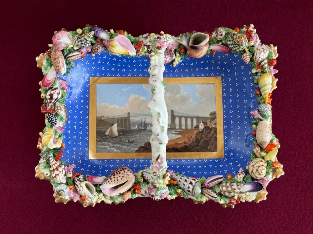 An antique rectangular porcelain basket with an overhead handle, decorated with three-dimensional applied shells along the rim and a hand-painted central view of the Menai Bridge on a cobalt blue dotted background.
