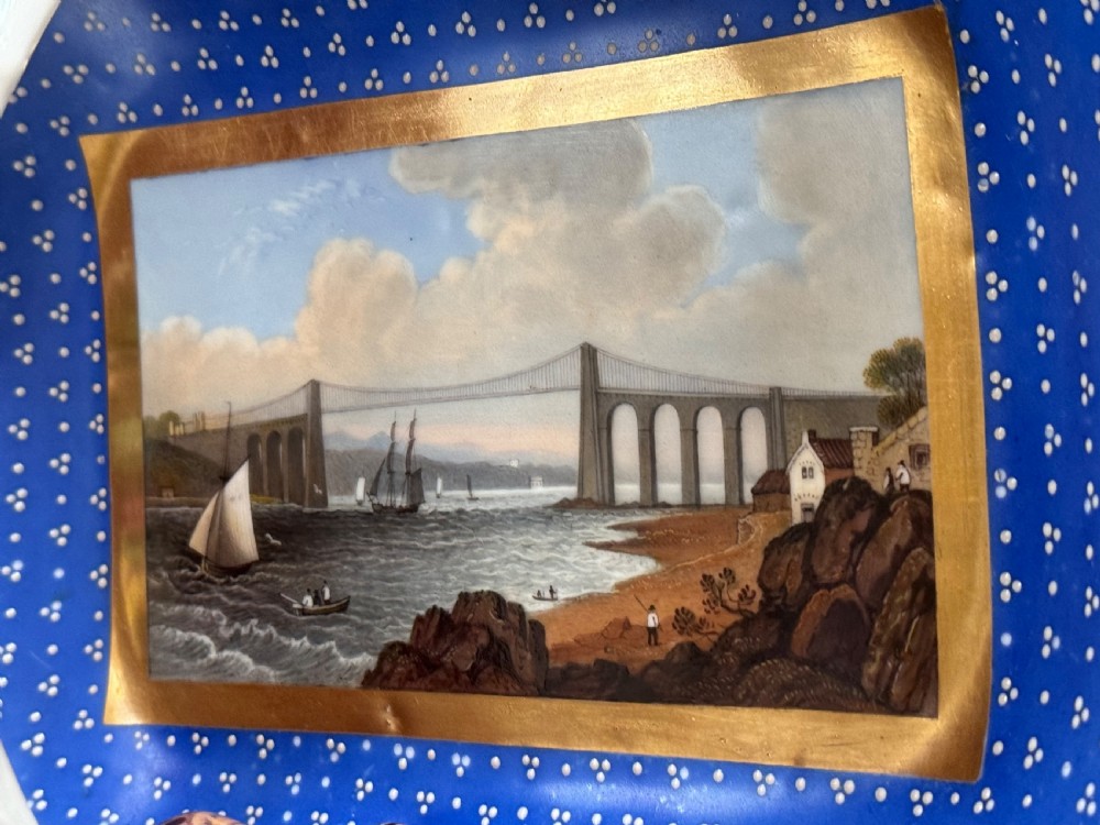 An antique rectangular porcelain basket with an overhead handle, decorated with three-dimensional applied shells along the rim and a hand-painted central view of the Menai Bridge on a cobalt blue dotted background. View II