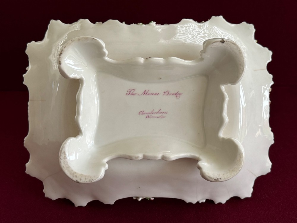 An antique rectangular porcelain basket with an overhead handle, decorated with three-dimensional applied shells along the rim and a hand-painted central view of the Menai Bridge on a cobalt blue dotted background. View IV