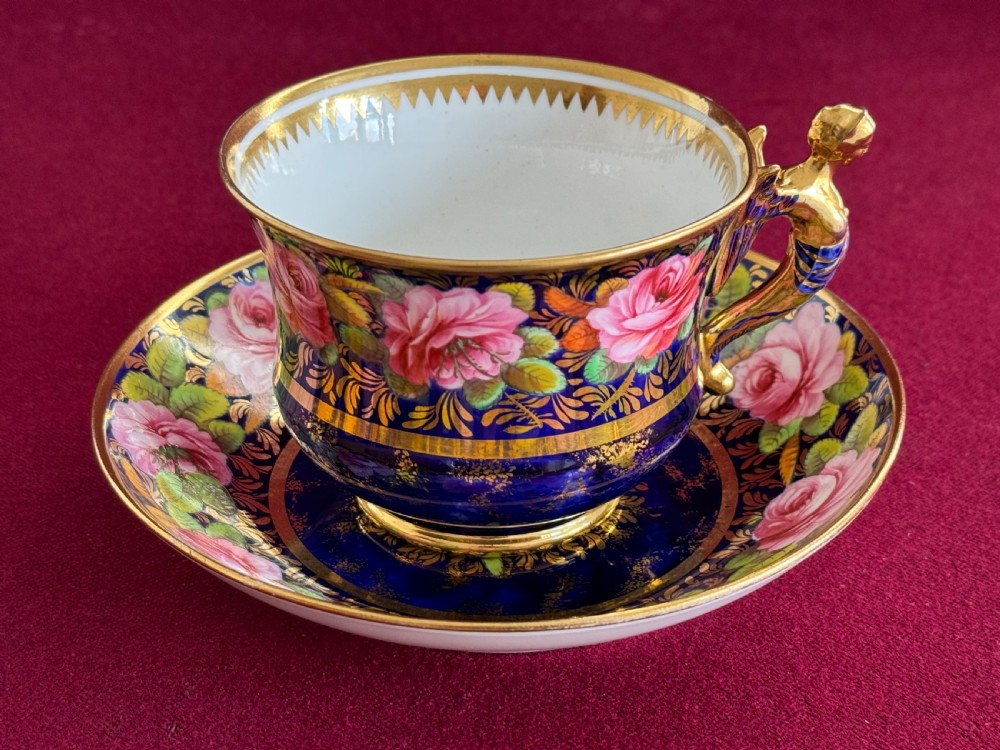 An antique Regency period Spode porcelain teacup and saucer featuring a cobalt blue background, hand-painted pink roses, gilded borders, and a gilded figural handle.
