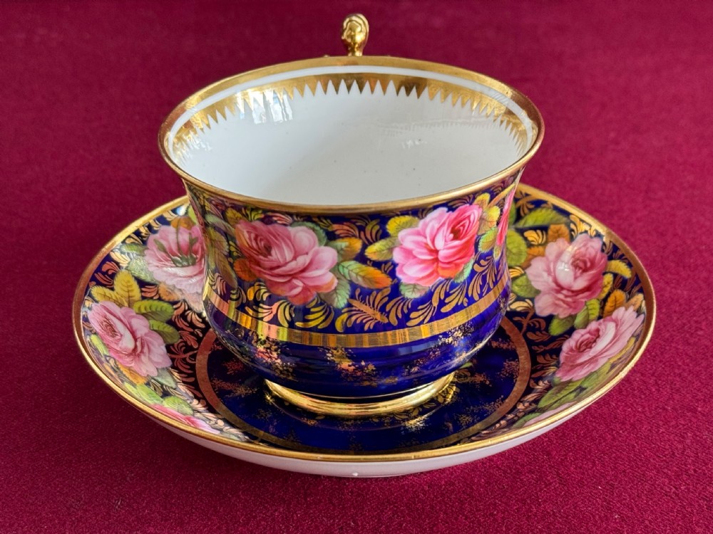 An antique Regency period Spode porcelain teacup and saucer featuring a cobalt blue background, hand-painted pink roses, gilded borders, and a gilded figural handle. View II