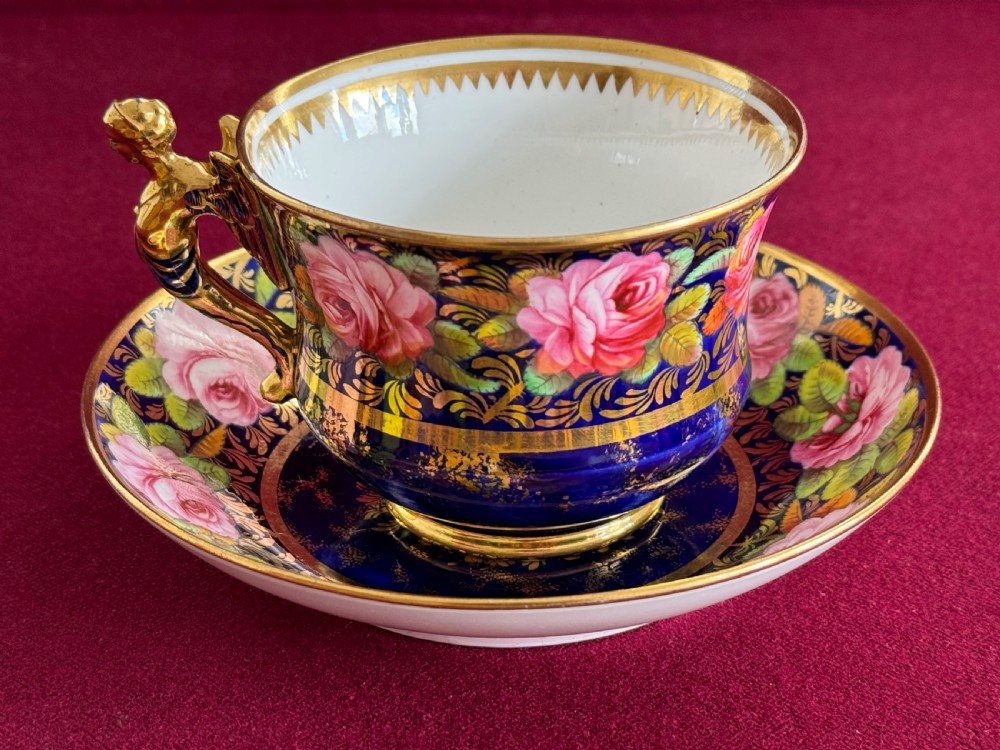 An antique Regency period Spode porcelain teacup and saucer featuring a cobalt blue background, hand-painted pink roses, gilded borders, and a gilded figural handle. View III