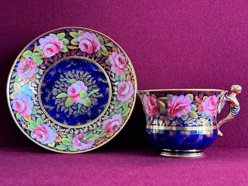 An antique Regency period Spode porcelain teacup and saucer featuring a cobalt blue background, hand-painted pink roses, gilded borders, and a gilded figural handle. View V