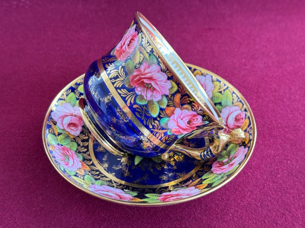 An antique Regency period Spode porcelain teacup and saucer featuring a cobalt blue background, hand-painted pink roses, gilded borders, and a gilded figural handle. View VI