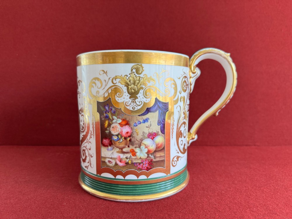 A cylindrical porcelain porter mug decorated with gold leaf scrollwork, a green reeded base, and a hand-painted floral still life panel.