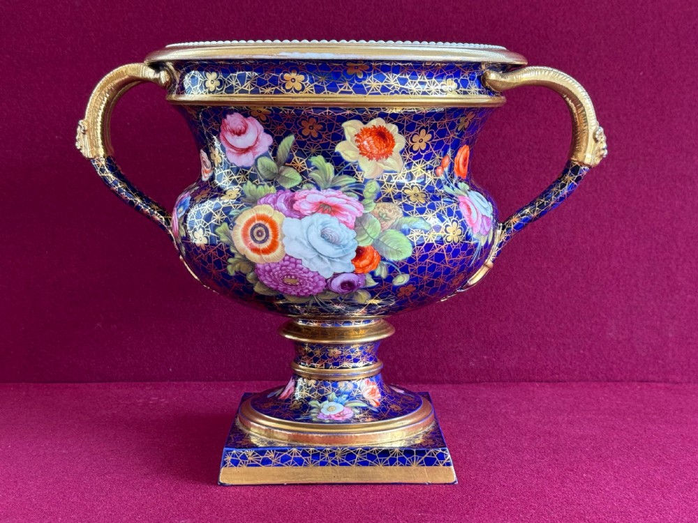 A cobalt blue Regency porcelain Warwick-style urn decorated with hand-painted floral bouquets, gold gilding, twin gilded handles, and a square pedestal base.