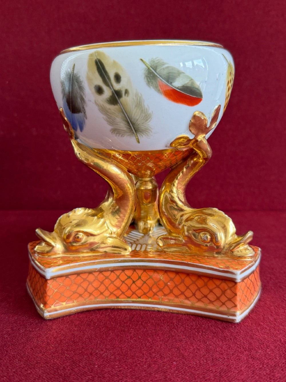 A Regency Chamberlain Worcester porcelain pastille burner decorated with hand-painted feathers and supported by three gilt dolphin legs on a patterned orange base.
