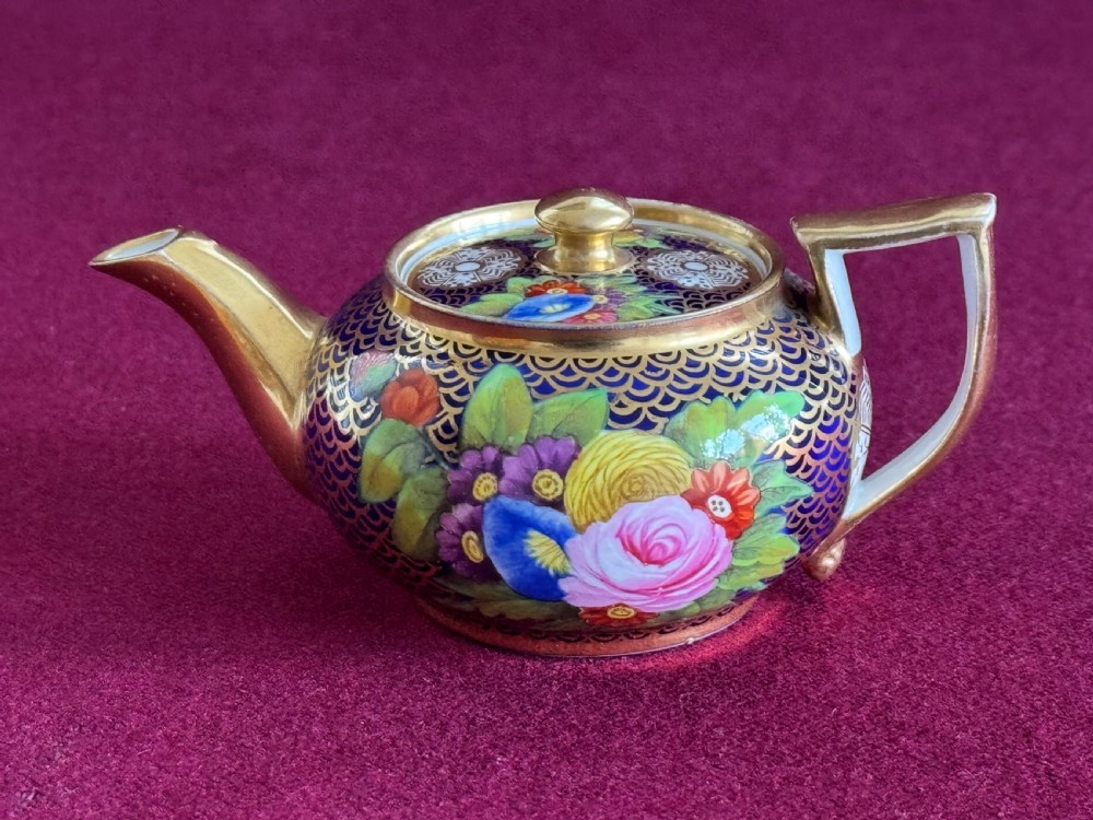 A miniature Regency porcelain teapot decorated with hand-painted floral panels on a cobalt blue ground enriched with gilded scale patterns.