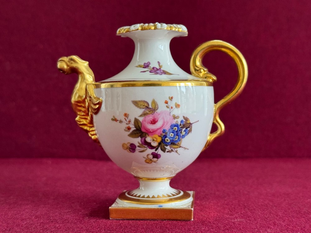 A small white porcelain rose water sprinkler with hand-painted floral panels, a gilded animal head spout, a gold scroll handle, and a square base.