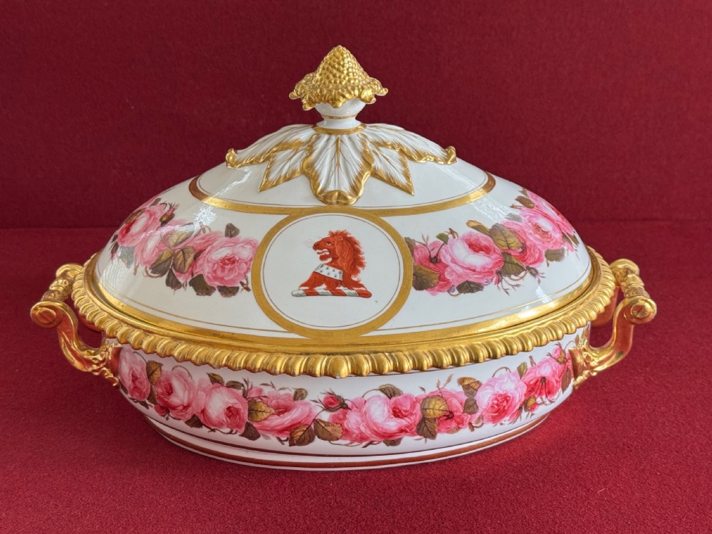 An oval Regency Worcester porcelain entree dish with a fitted lid, featuring hand-painted pink rose garlands, gilt gadrooned rims, twin gilded handles, a heraldic lion crest, and a gold leaf-and-pineapple finial.