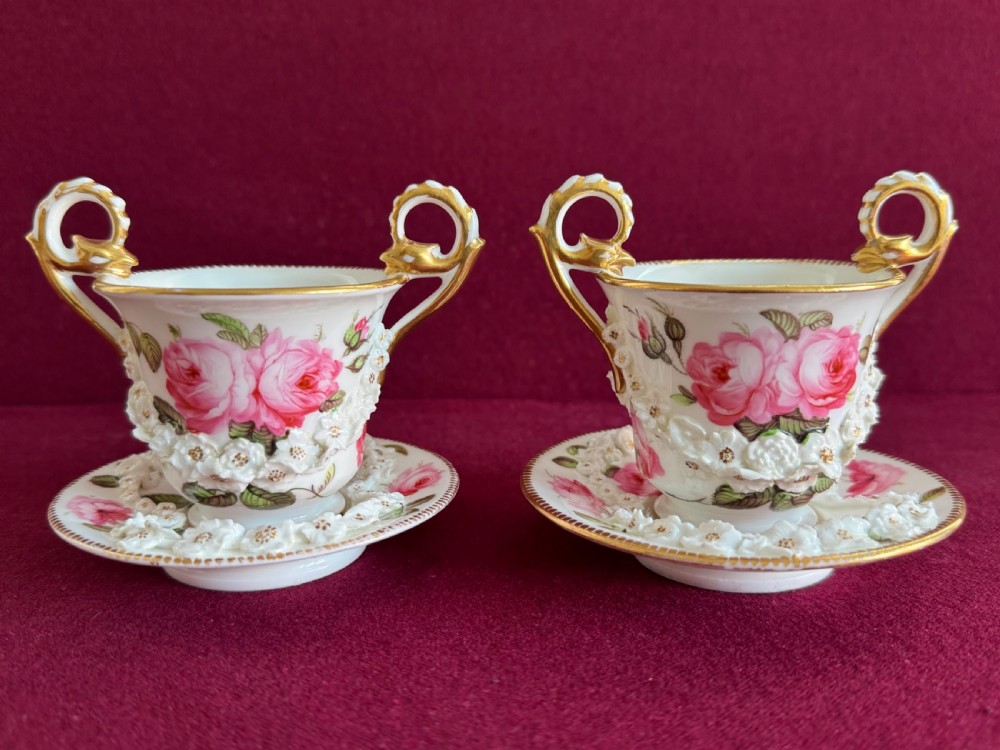 A pair of white Regency porcelain cabinet cups and saucers decorated with hand-painted pink roses, applied porcelain flowers, and ornate gilt handles.