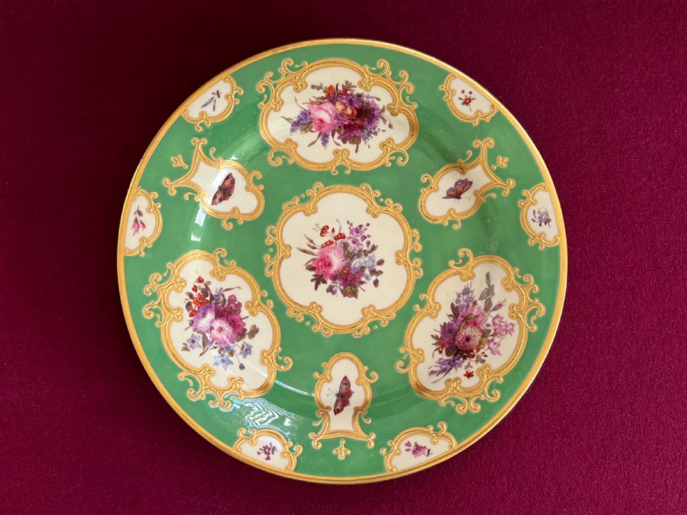 A circular green porcelain plate featuring hand-painted floral cartouches surrounded by gilded rococo scrollwork and central floral and butterfly vignettes.
