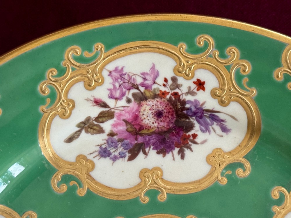 A circular green porcelain plate featuring hand-painted floral cartouches surrounded by gilded rococo scrollwork and central floral and butterfly vignettes. View V