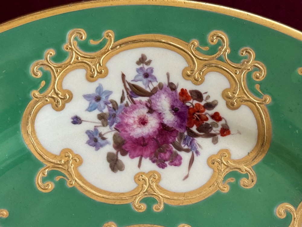 A circular green porcelain plate featuring hand-painted floral cartouches surrounded by gilded rococo scrollwork and central floral and butterfly vignettes. View VI