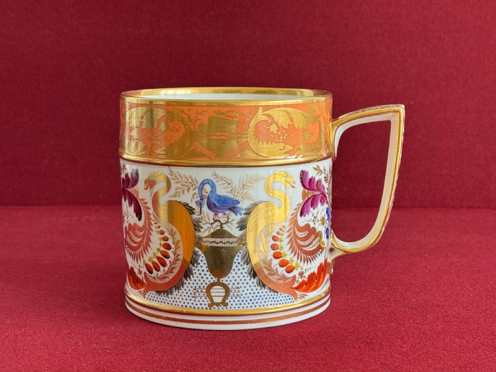 A Regency Derby porcelain porter mug featuring hand painted bird motifs, an orange gilded upper border, and an angular handle.