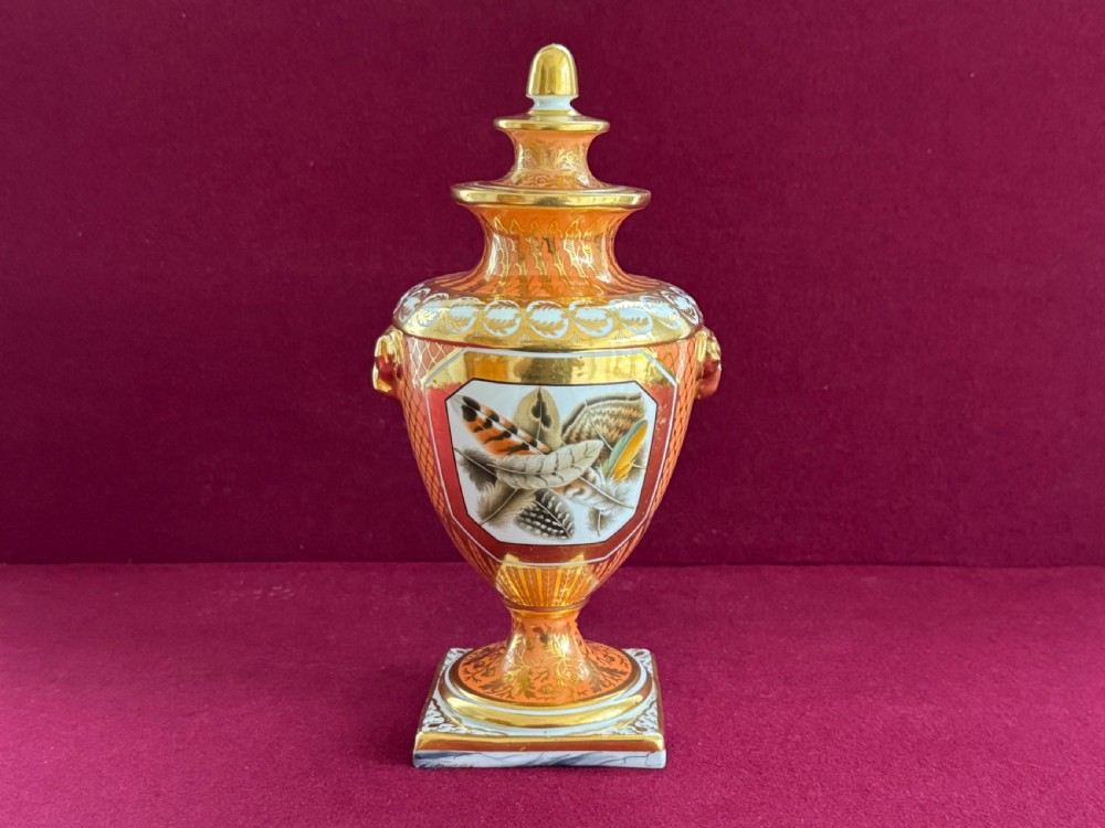 A Regency Chamberlain Worcester porcelain urn-shaped vase with a lid, featuring an orange pattern ground, gilded accents, side mask handles, and a painted feather panel on a square base.