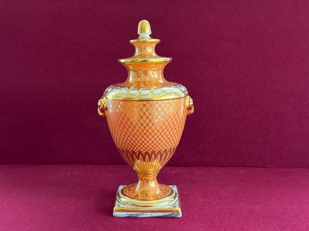 A Regency Chamberlain Worcester porcelain urn-shaped vase with a lid, featuring an orange pattern ground, gilded accents, side mask handles, and a painted feather panel on a square base. View III