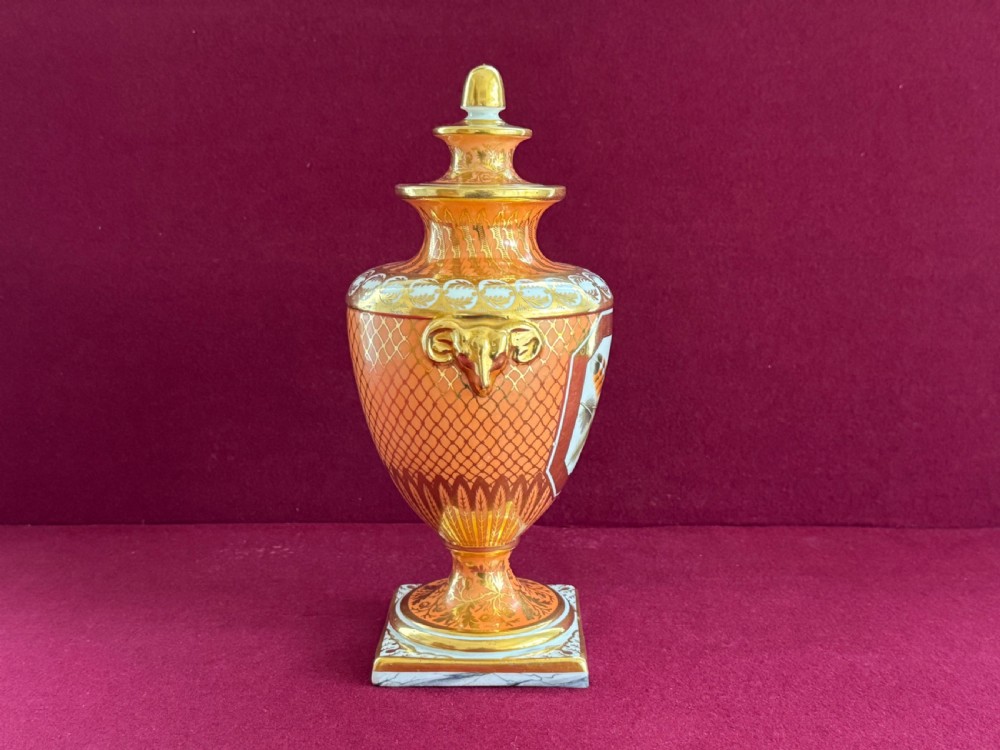 A Regency Chamberlain Worcester porcelain urn-shaped vase with a lid, featuring an orange pattern ground, gilded accents, side mask handles, and a painted feather panel on a square base. View IV