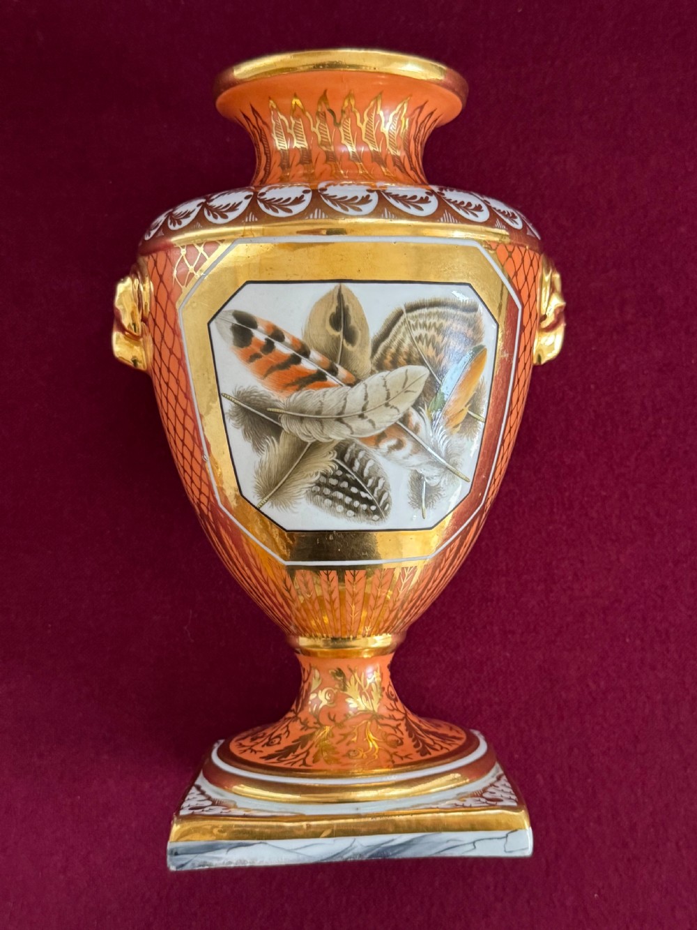 A Regency Chamberlain Worcester porcelain urn-shaped vase with a lid, featuring an orange pattern ground, gilded accents, side mask handles, and a painted feather panel on a square base. View VI