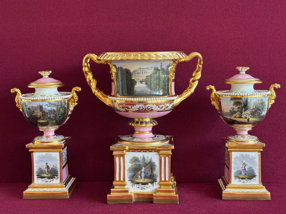 A three-piece Flight, Barr and Barr Worcester porcelain garniture set with hand-painted landscape scenes, pink ground borders, and gilded scroll handles resting on square pedestal bases.