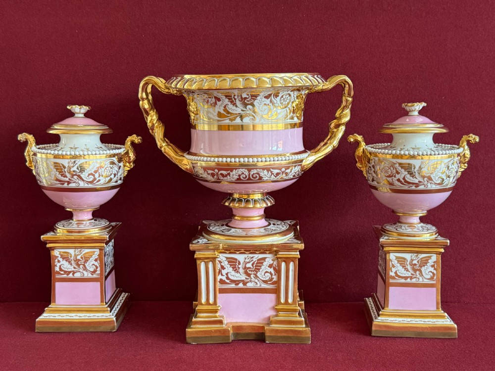 A three-piece Flight, Barr and Barr Worcester porcelain garniture set with hand-painted landscape scenes, pink ground borders, and gilded scroll handles resting on square pedestal bases. View VIII