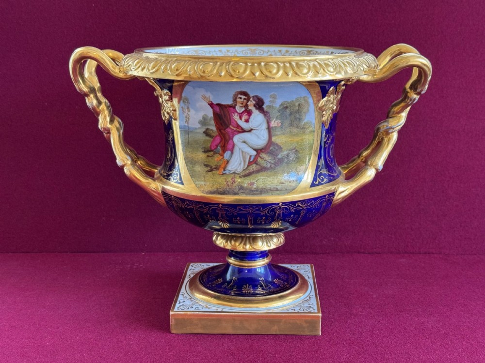 A Regency Flight, Barr and Barr Worcester cobalt blue porcelain Warwick urn featuring twin gilt vine-twisted handles, intricate gilt tooling, and a central hand-painted panel depicting two figures in a landscape.