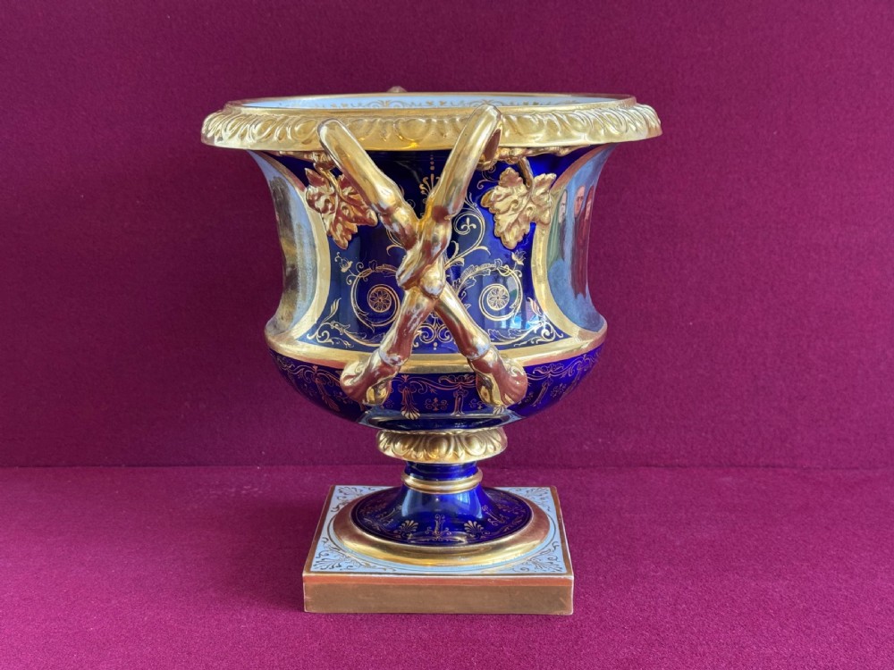 A Regency Flight, Barr and Barr Worcester cobalt blue porcelain Warwick urn featuring twin gilt vine-twisted handles, intricate gilt tooling, and a central hand-painted panel depicting two figures in a landscape. View II
