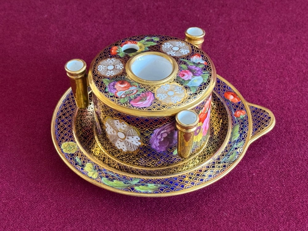 An early 19th-century circular English porcelain inkstand featuring gold scale gilding on a dark blue ground, floral reserves, and three cylindrical quill holders.