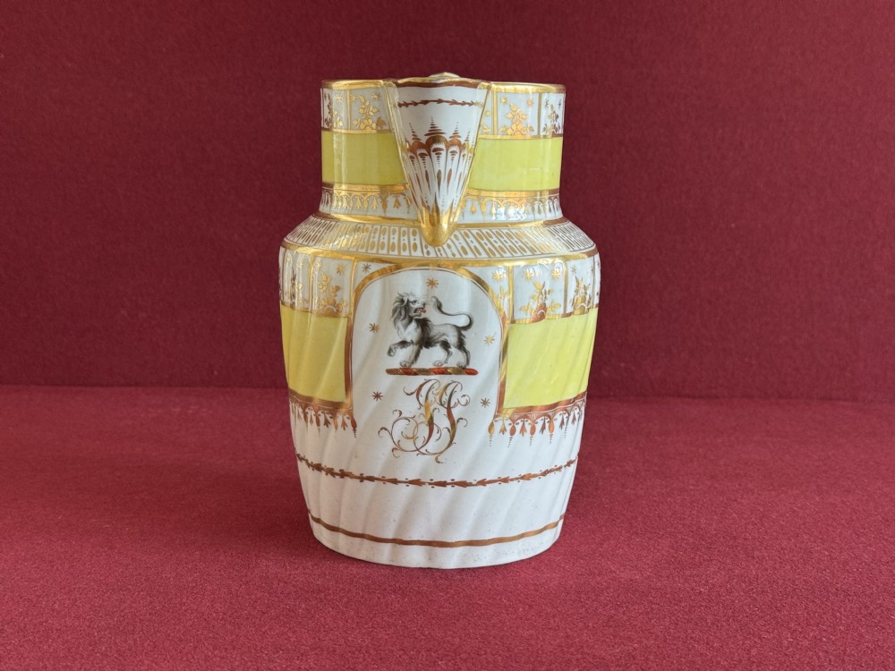 A white and yellow antique porcelain jug decorated with gilt detailing, spiral fluting, and a central armorial crest featuring a painted lion.