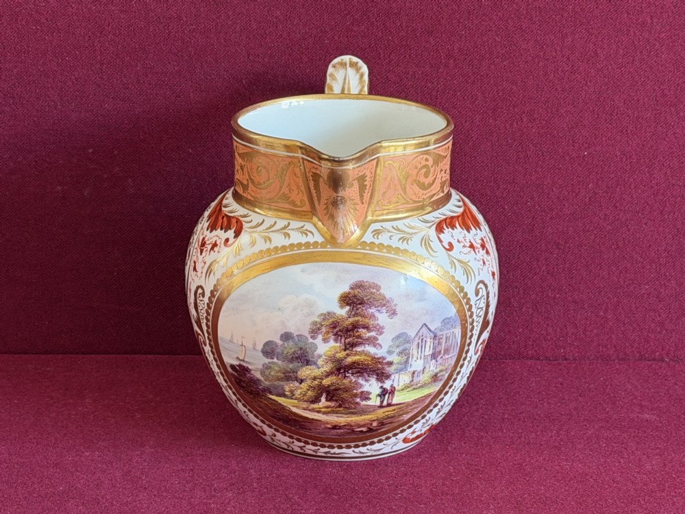 A Regency era Derby porcelain jug featuring a hand-painted landscape panel framed by gold leaf cartouche details and coral-tinted scrollwork.