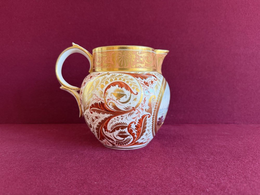 A Regency era Derby porcelain jug featuring a hand-painted landscape panel framed by gold leaf cartouche details and coral-tinted scrollwork. View VI